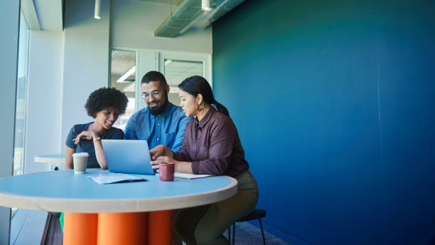 Diverse group of a smiling young businesspeople working together on a laptop during a meeting around a table in an office lounge